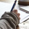 person writing on brown wooden table near white ceramic mug
