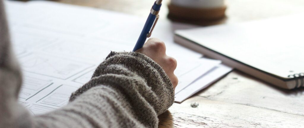 person writing on brown wooden table near white ceramic mug