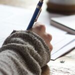 person writing on brown wooden table near white ceramic mug